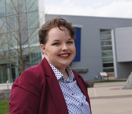 Picture of a short-haired individual wearing a maroon suit partially angled toward the viewer. 
      They are wearing rose pink lipstick and a toothy smile. The button down under the jacket is white, 
      patterned with indeterminate circular shapes. Behind them is a gray building with a wall made of 
      windows and a blue accent. A leafless tree is visible to the side behind their head.
